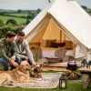 Two men with two dogs sitting outside a white canvas glamping tent in a rural countryside setting.